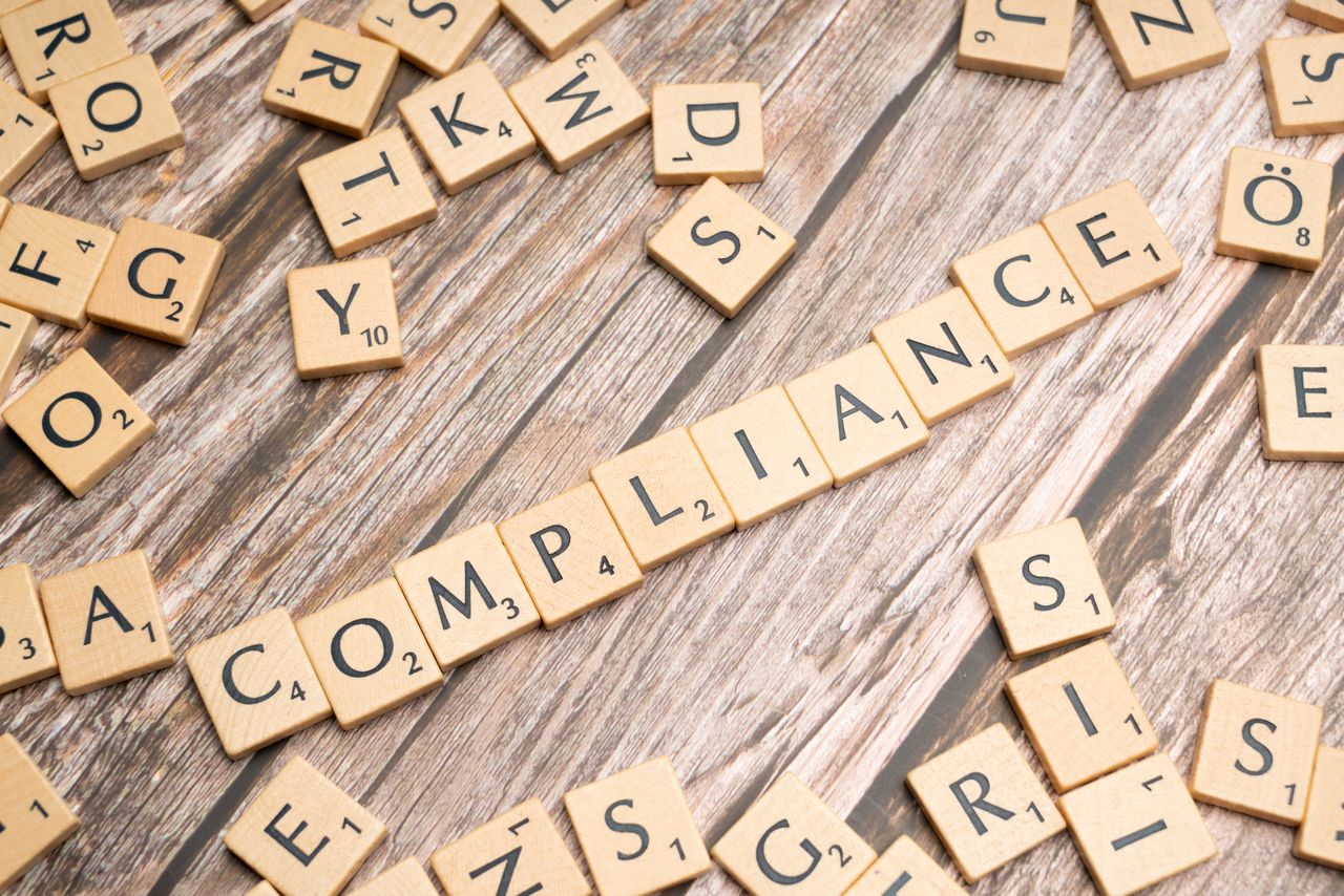 hero-img-02 Wooden letter tiles forming the word 'COMPLIANCE' on a rustic wooden background.
